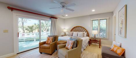 A bedroom with a bed, two armchairs, a nightstand, a ceiling fan, and a large window with a view of palm trees.