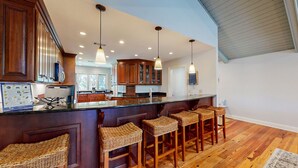 A modern kitchen with wooden cabinets, a central island with bar stools, pendant lighting, and a view of the dining area. - Golf course and lagoon views! (Hilton Head)
