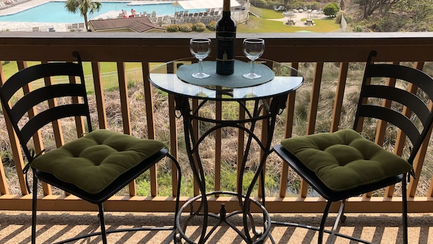 En balkon med to polstrede stole, et glastabel og en flaske vin. En pool og strand ses i baggrunden.