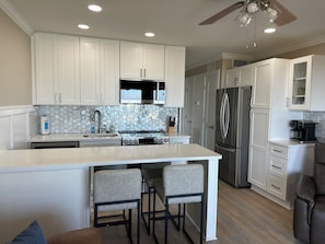 A modern kitchen with white cabinets, a central island with bar stools, stainless steel appliances, and a ceiling fan.