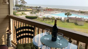 En balkon med et bord, to vinglas og en vinflaske. Udsigten inkluderer en swimmingpool, en strand og et resortområde.