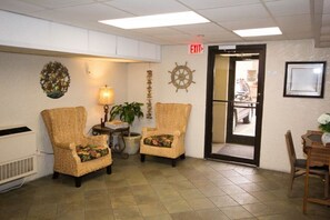 A lobby with wicker chairs, a lamp, a ship's wheel decoration, and an exit sign.