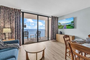 A room with a balcony, a dining table set, a TV on the wall, and a view of the sea.