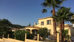 A Mediterranean-style villa with a stone wall, palm trees, and a clear blue sky.