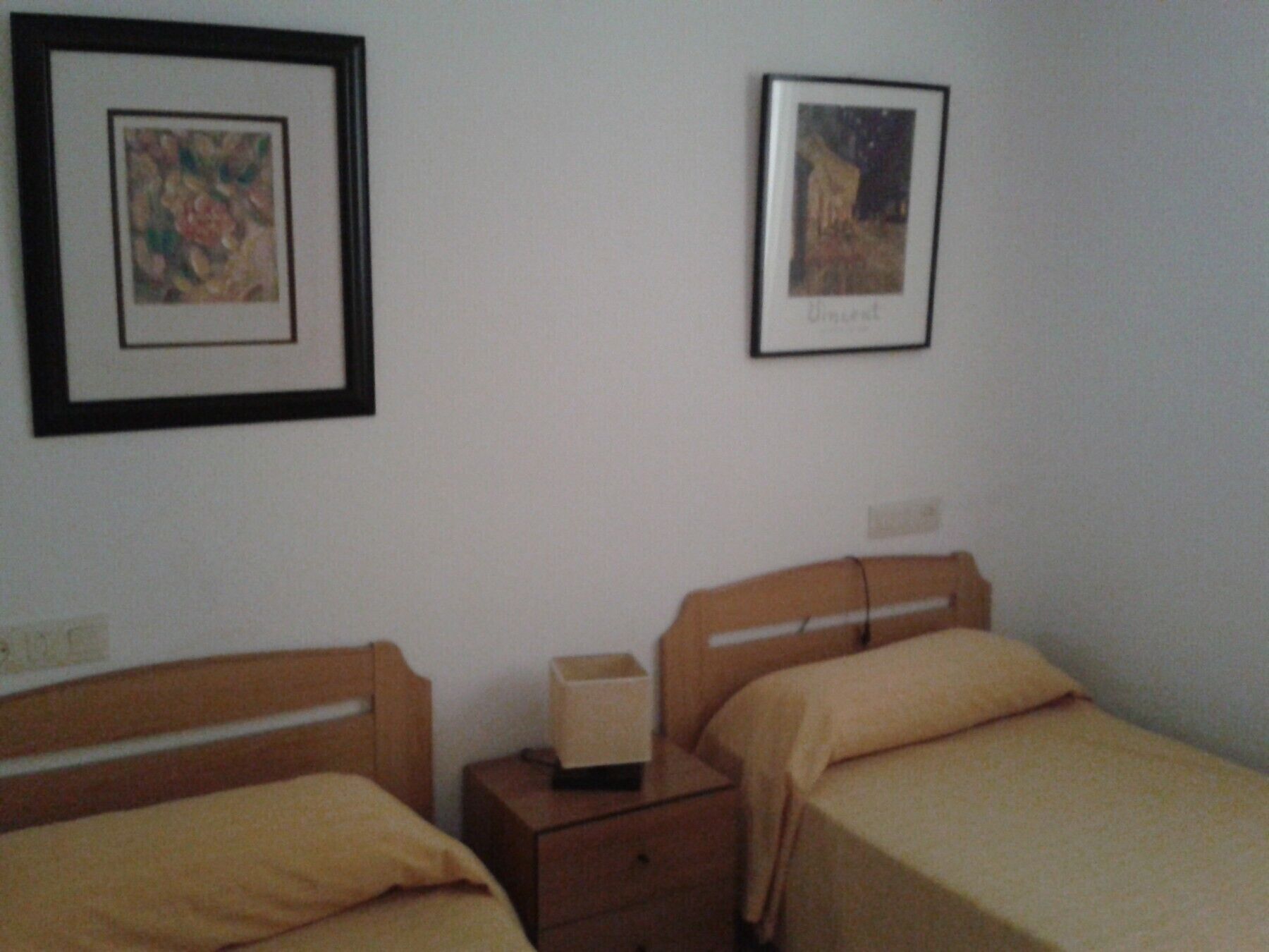 A small, simple room with two single beds, a nightstand, and two framed pictures on the wall.
