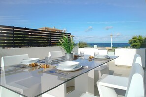 A modern outdoor dining area with a glass table, white chairs, and a view of the ocean. - Atzur Plus 177 modern villa with private pool, air-conditioning, gym and BBQ (Son Serra de Marina)