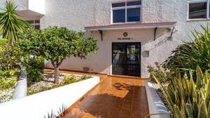 A building with a white exterior, a black door, and a small window above the door. There is a tree and some plants in front of the building.