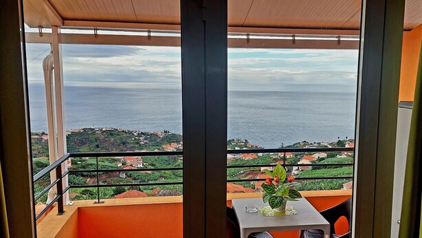 A balcony with a table and chairs, overlooking a coastal landscape with houses and greenery. - Villa E Apartment (Ponta do Sol)