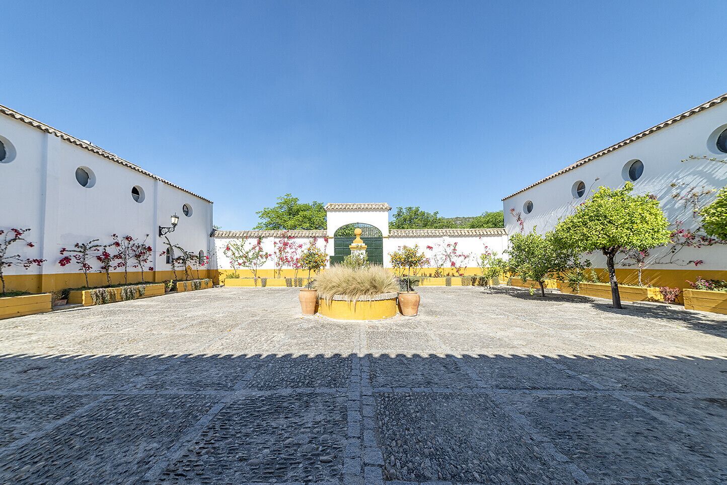 Ein Innenhof mit einem zentralen Brunnen, umgeben von weißen Gebäuden mit gelbem Verzierband und grünen Türen.