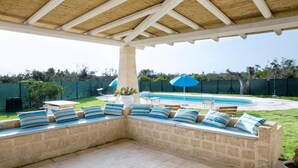 Ein überdachter Außenbereich mit Steinmauern und blau-weiß gestreiften Kissen, mit Blick auf einen Swimmingpool und einen eingezäunten Garten.