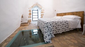 Ein Schlafzimmer mit Steinboden, einem Bett mit gemustertem Überwurf, einem Holz-Kopfteil und einem kleinen Fenster.