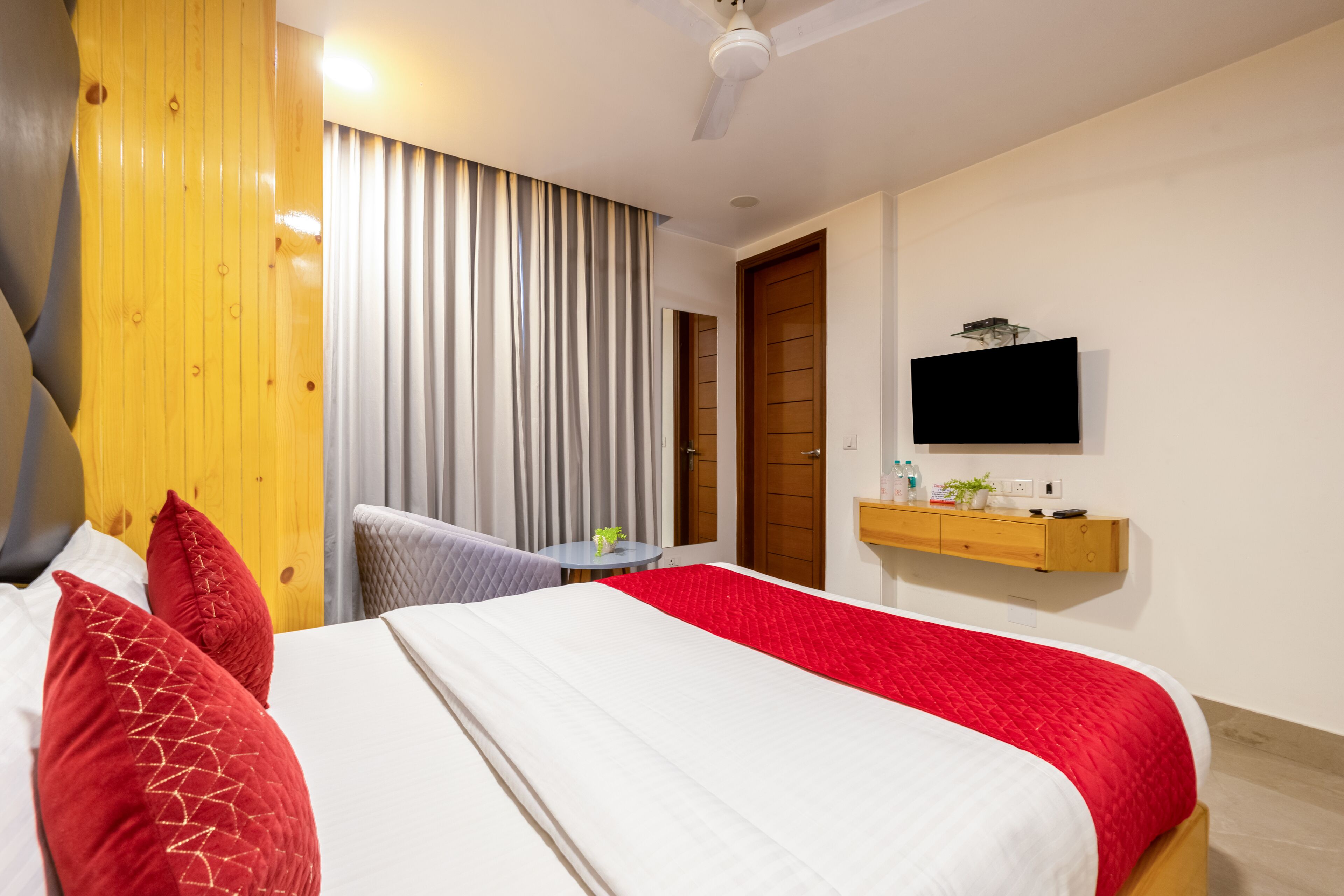 A hotel room with a bed, a TV, a small table with a plant, and a wooden shelf with toiletries.