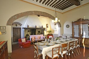 Un coin repas avec une longue table dressée pour le repas, un canapé rouge et une cheminée ornée de pots suspendus.