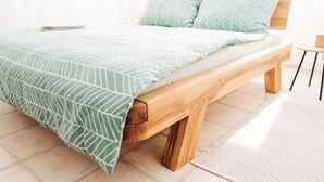 Una cama con colcha y almohadas estampadas con motivos de hojas, un banco de madera y una alfombra con diseño sobre suelo de baldosas.