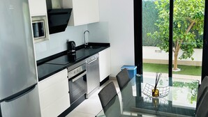 Eine moderne Küche mit weißen Schränken, einem Edelstahlkühlschrank und einem eingebauten Backofen. Es gibt eine Essecke mit einem Glastisch und Stühlen, und durch große Fenster ist ein Gartenblick möglich.