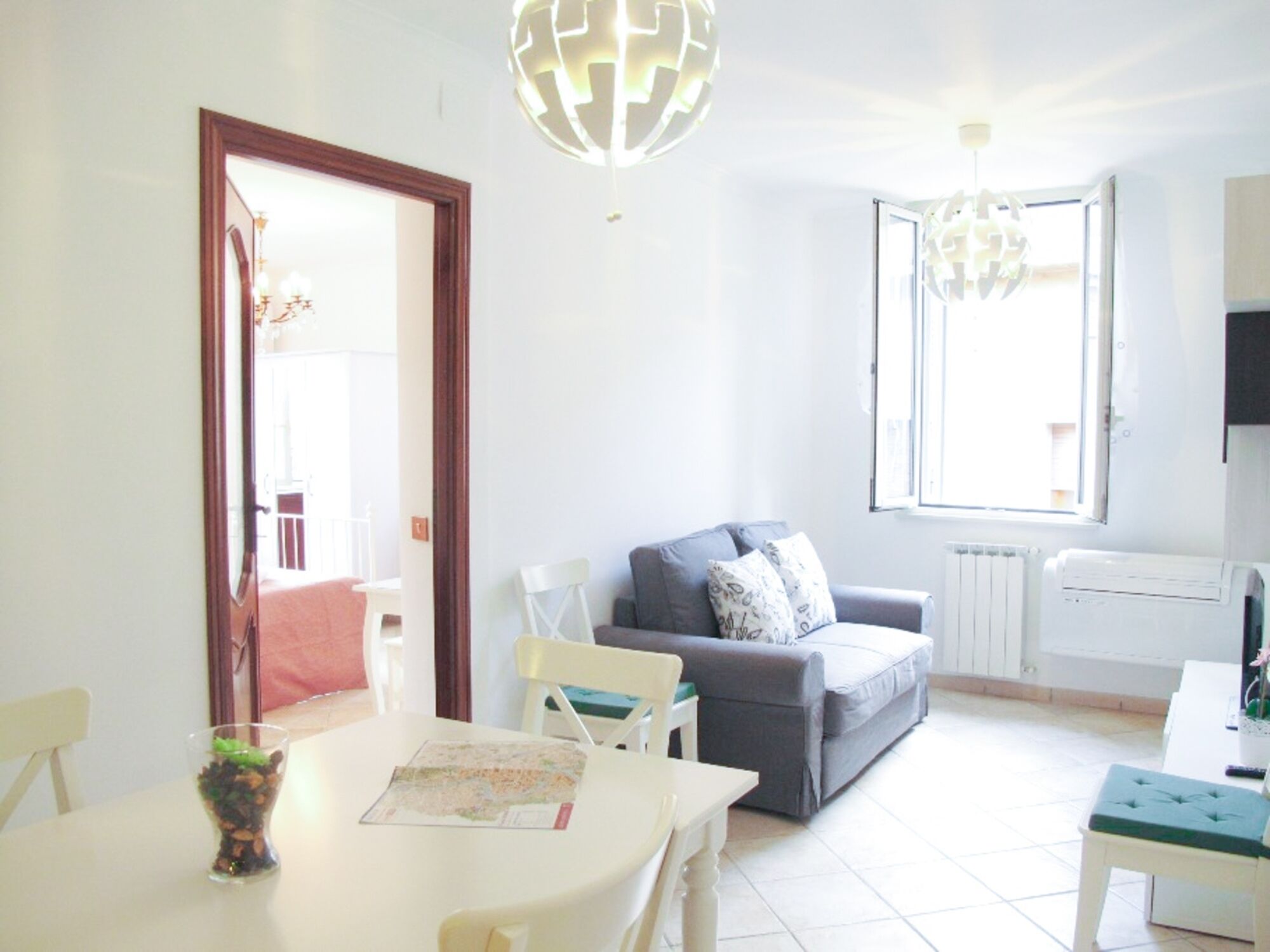 A bright living room with a gray sofa, a white dining table, and a glass coffee table. There are two chairs, a white radiator, and a window with a view.