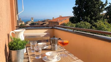 Un balcone con un tavolo apparecchiato per due persone, una pianta in vaso, una moka e un bicchiere di bevanda.