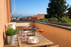 Un balcón con una mesa lista para dos personas, una planta en maceta, una cafetera y un vaso de bebida.