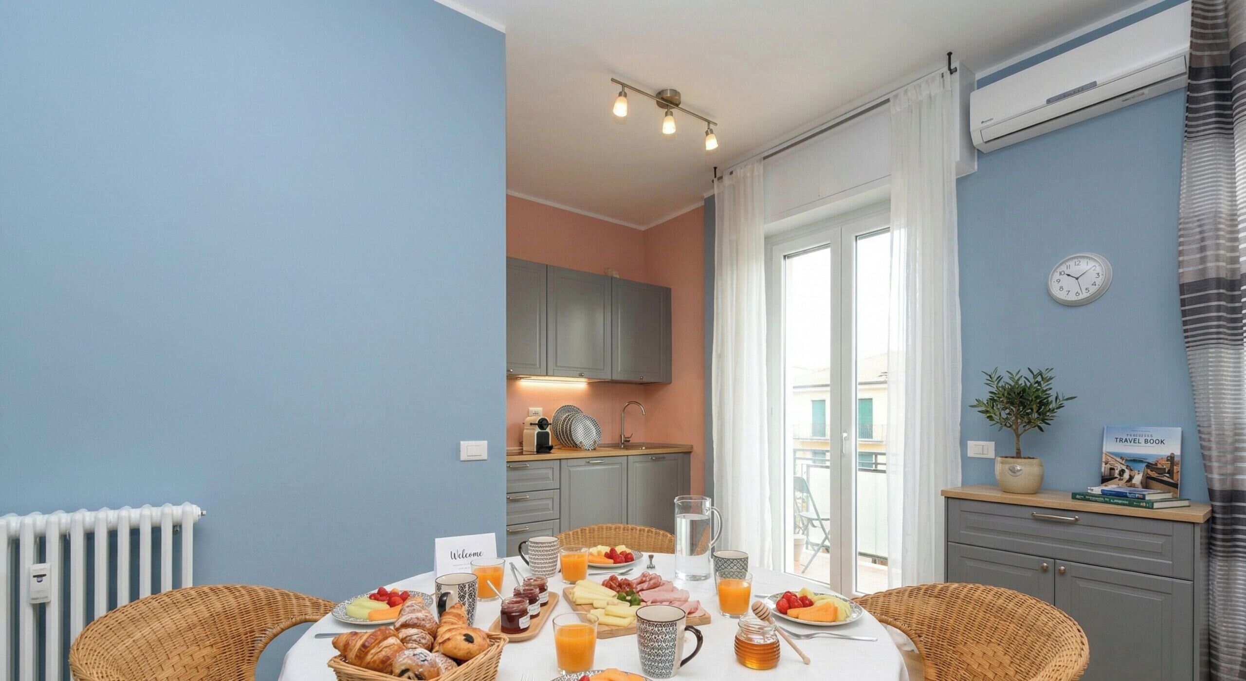 Una colazione servita con un tavolo, sedie in vimini, una cucina sullo sfondo e un orologio al muro.