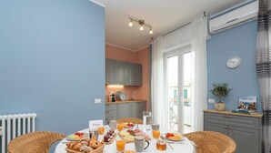 Desayuno montado sobre una mesa, con sillas de mimbre, una cocina al fondo y un reloj en la pared.
