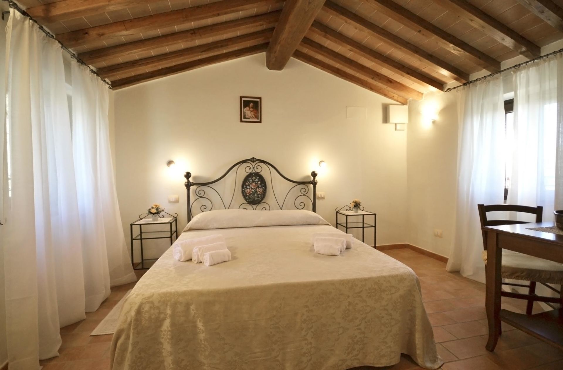 Un dormitorio con una cama, dos mesitas de noche, una silla y una pequeña mesa con una lámpara.