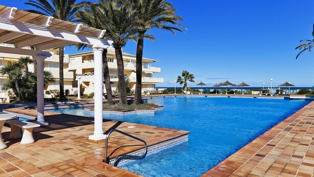A swimming pool with a tiled deck, palm trees, and a building in the background.