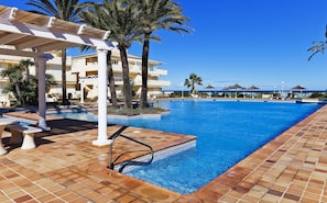 A swimming pool with a tiled deck, palm trees, and a building in the background. - 106 Playa Dorada J-1 (Denia)
