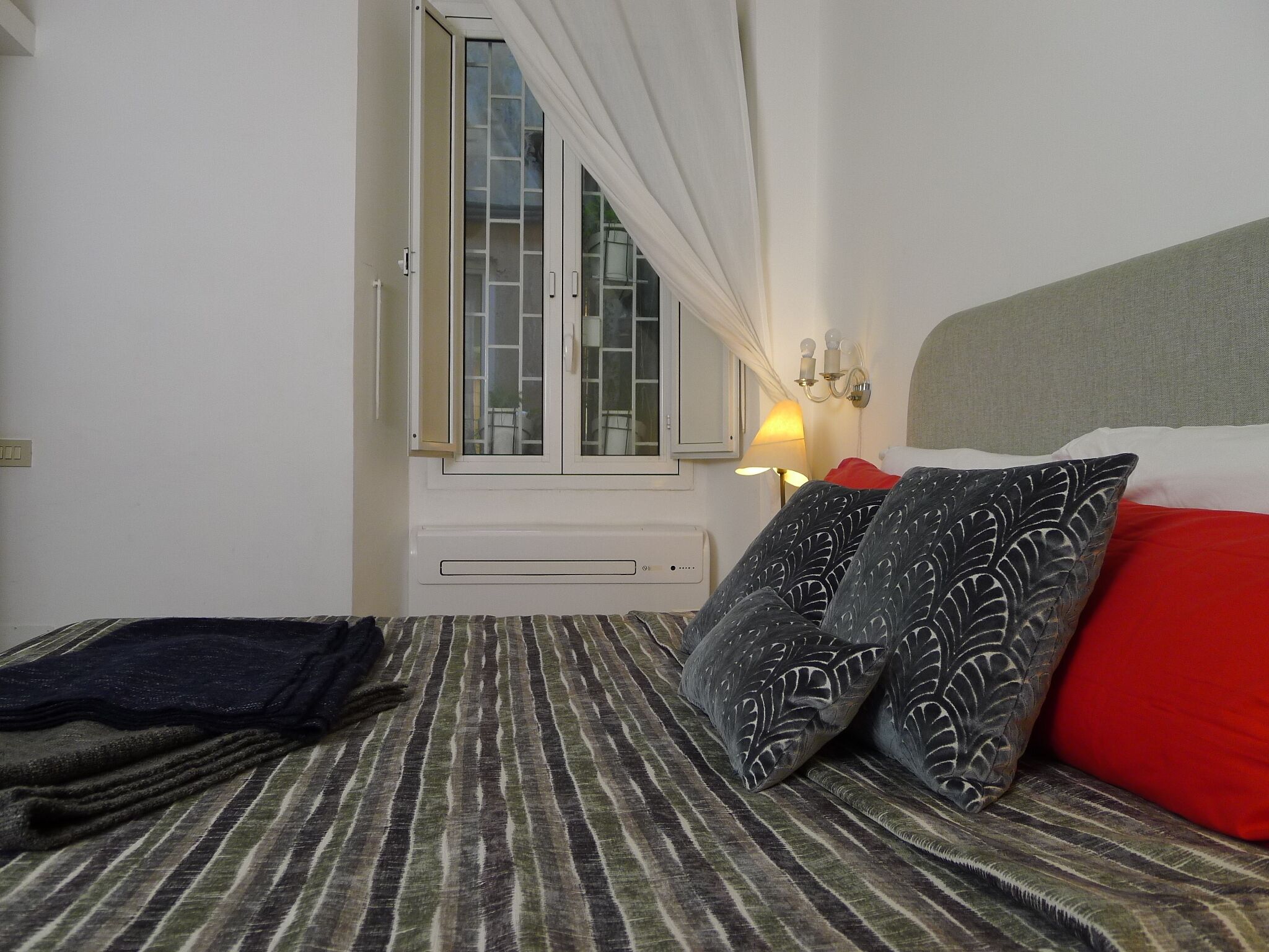 Quarto de hotel com cama, janela com cortinas, luminária na parede e travesseiro vermelho.