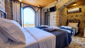 A bedroom with two beds, a stone wall, a television, and a view of the sea.