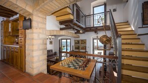 Un vaste espace de vie avec une grande table en bois, prête pour une partie, un escalier doté d’une rampe en fer forgé et un mur en pierre.