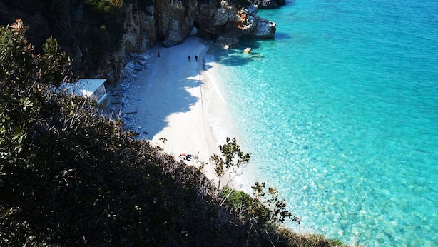 A coastal beach with clear turquoise water, rocky cliffs, and sparse vegetation.