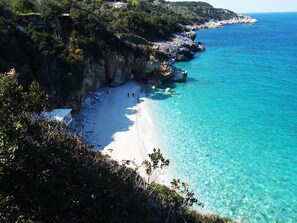 A coastal beach with clear turquoise water, rocky cliffs, and sparse vegetation.