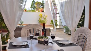 Un área de comedor al aire libre con una mesa dispuesta para cuatro personas, rodeada de cortinas blancas, sillas de mimbre y plantas en macetas.