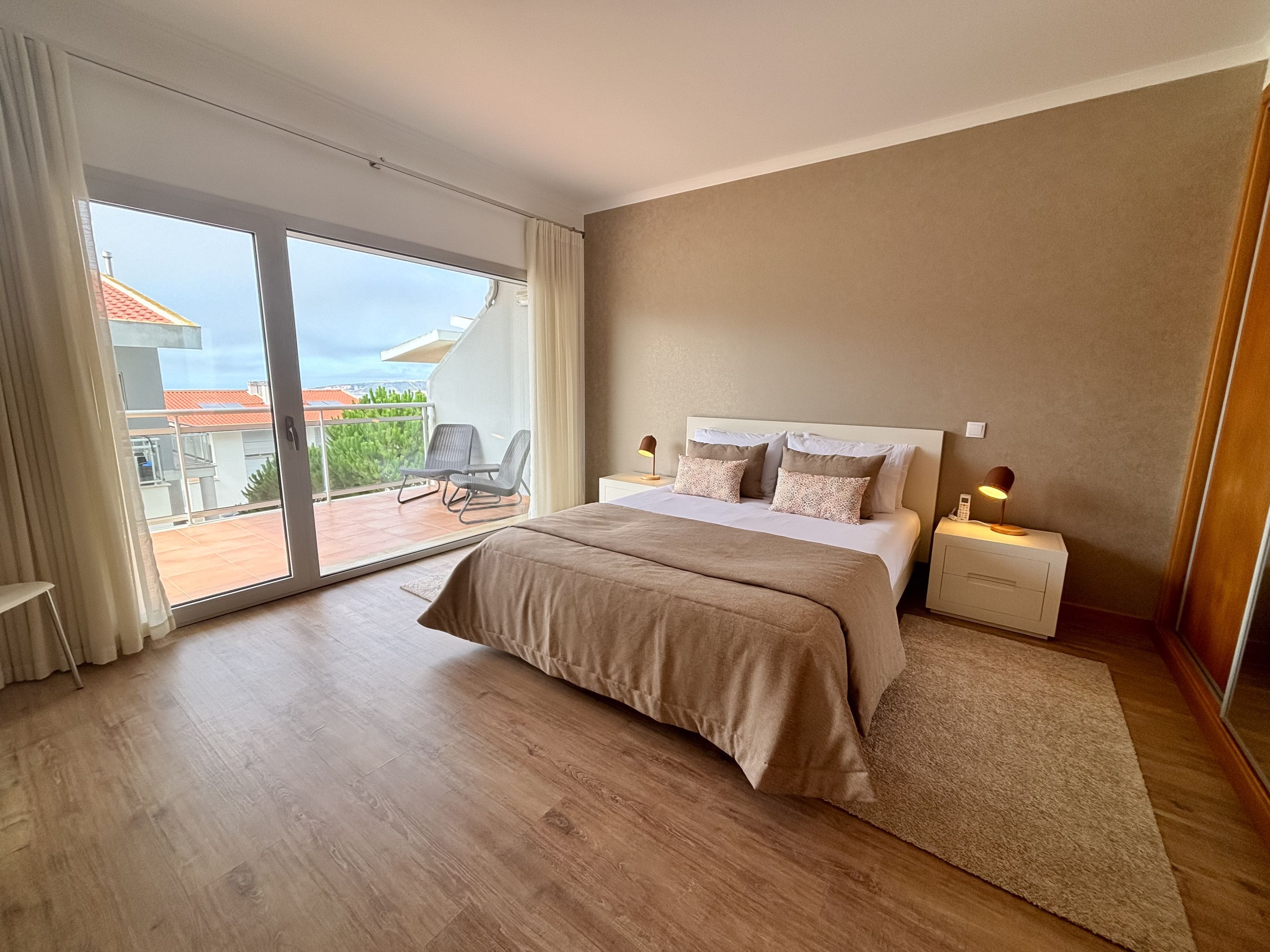 A bedroom with a large sliding glass door leading to a balcony, a bed with a headboard, bedside tables, and a view of buildings.