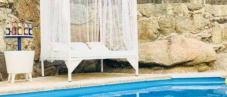 Una cama con dosel blanco junto a una piscina, con un muro de piedra de fondo.