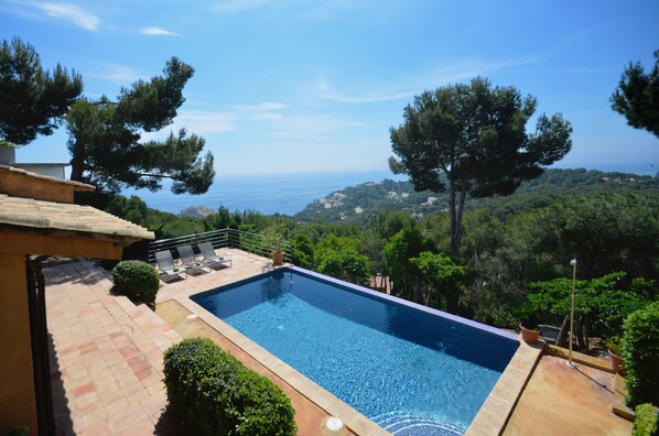 A swimming pool with a view of the sea, surrounded by trees and a patio area.