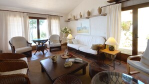 A cozy living room with a white sofa, two armchairs, a coffee table, and a small side table with a lamp. There is a window with curtains, a painting on the wall, and a wooden shelf above the sofa. - Holiday rental villa with swimming pool in Tamariu, Tamariu (Tamariu)
