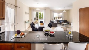 A modern kitchen with a black countertop, white chairs, and a dining table. There is a view of a balcony with outdoor furniture and greenery.