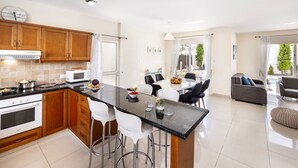 A modern kitchen with wooden cabinets, a black countertop, and stainless steel appliances. There is a dining area with a round table and chairs, and a living area with a sofa and a coffee table.