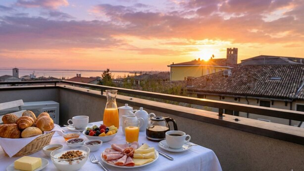 Morgenmad på en balkon med udsigt over byen i solnedgang, med croissanter, frugt, ost og kaffe.