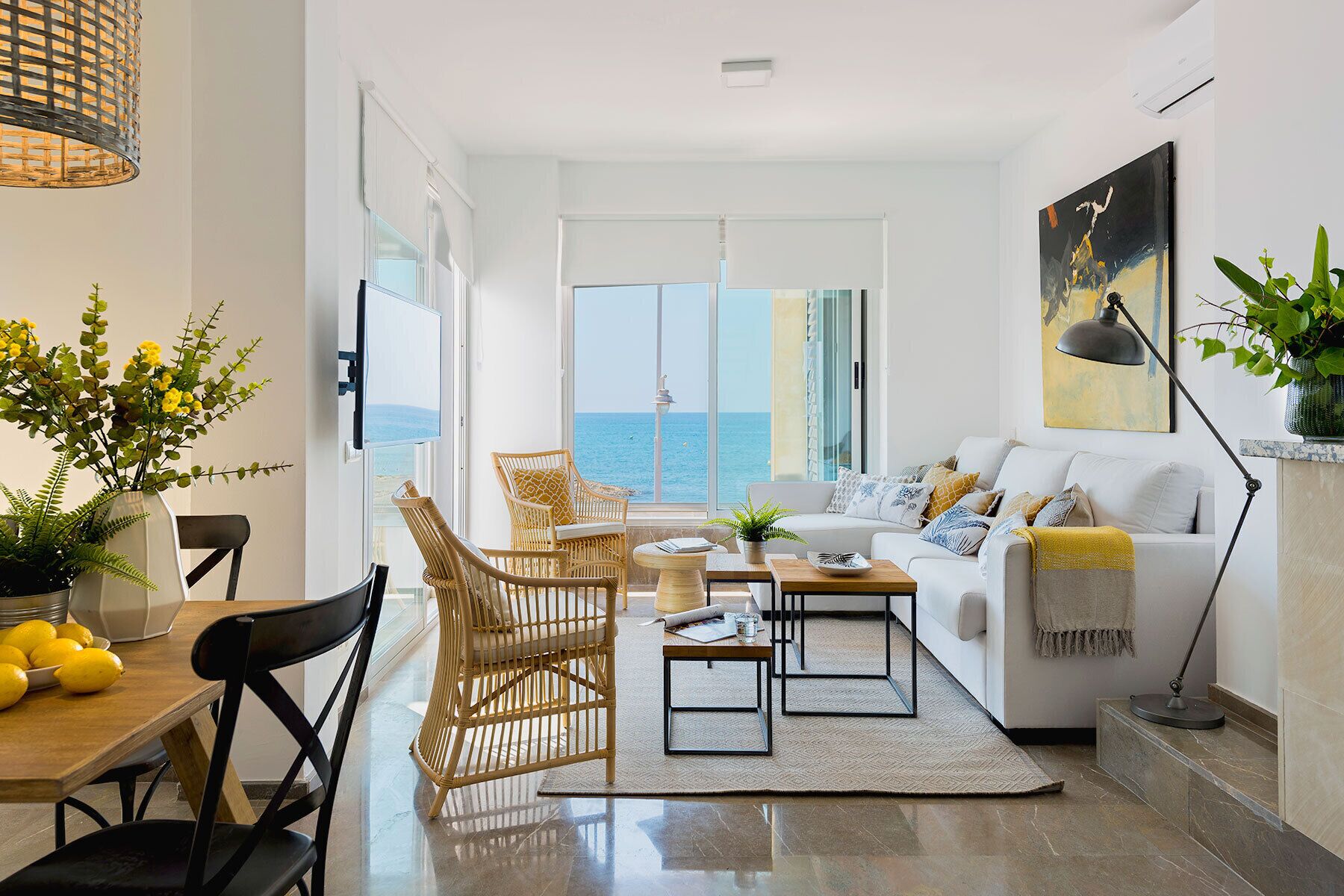 A modern living room with a white sofa, wicker chairs, a dining table with lemons, and a view of the sea.