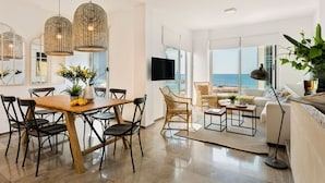 A modern living room with a large window, a dining area with a wooden table, wicker chairs, and a view of the sea.