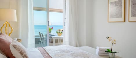 A hotel room with a bed, a table with flowers, a view of the sea, and two framed pictures on the wall.