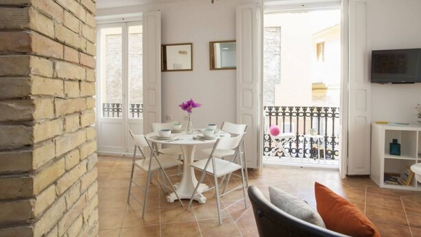 A dining area with a white table and chairs, a vase with flowers, and a balcony with a black railing.