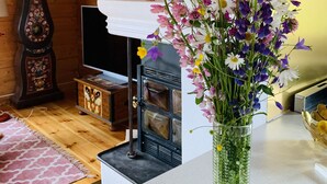 Een knus vertrek met een houten plafond, een antieke klok, een open haard en een vaas bloemen op een witte tafel.
