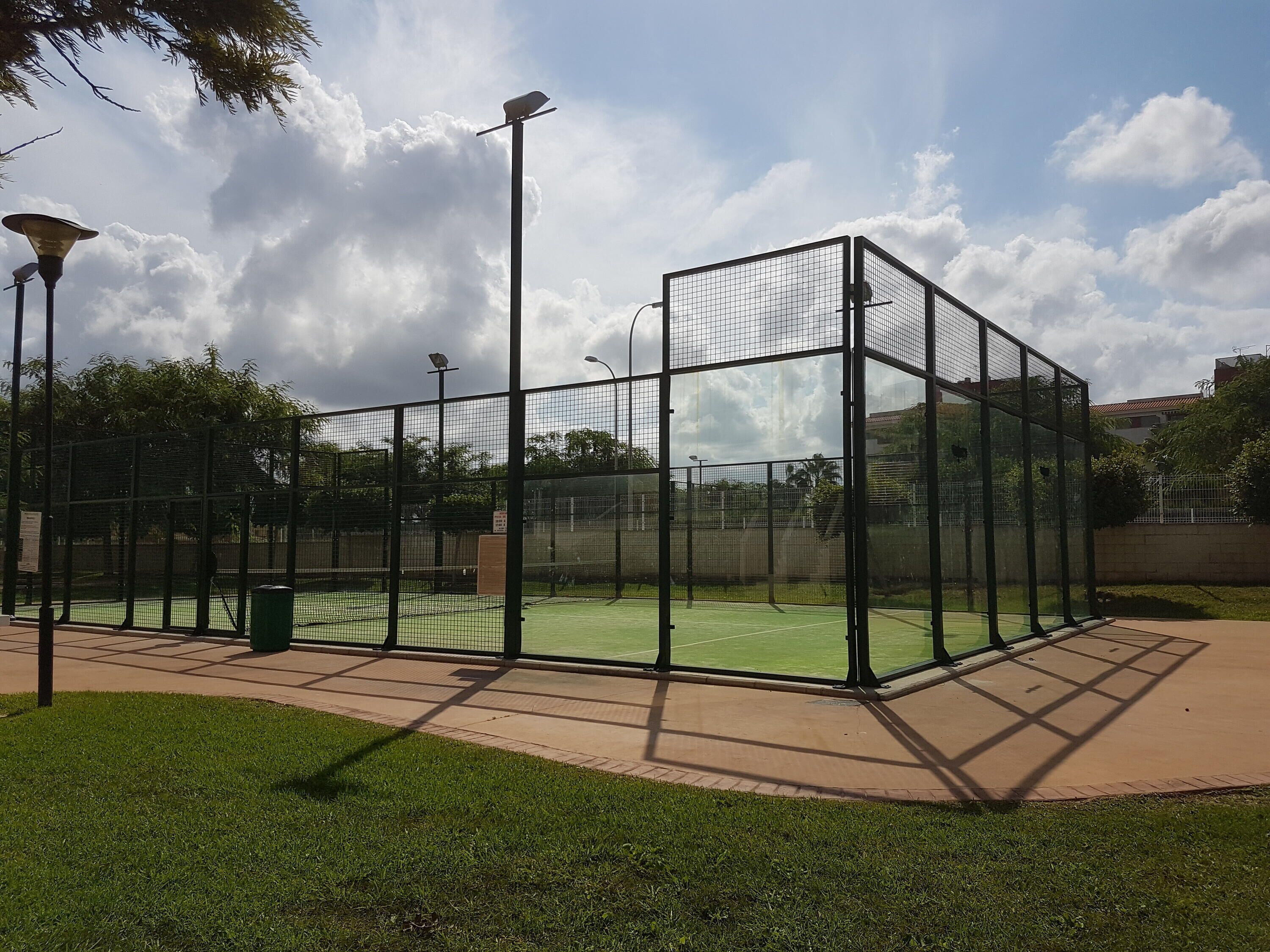 Ein Tennisplatz, umgeben von einem schwarzen Metallzaun und einer begrünten Oberfläche, mit einem gepflasterten Weg und Straßenlaternen.