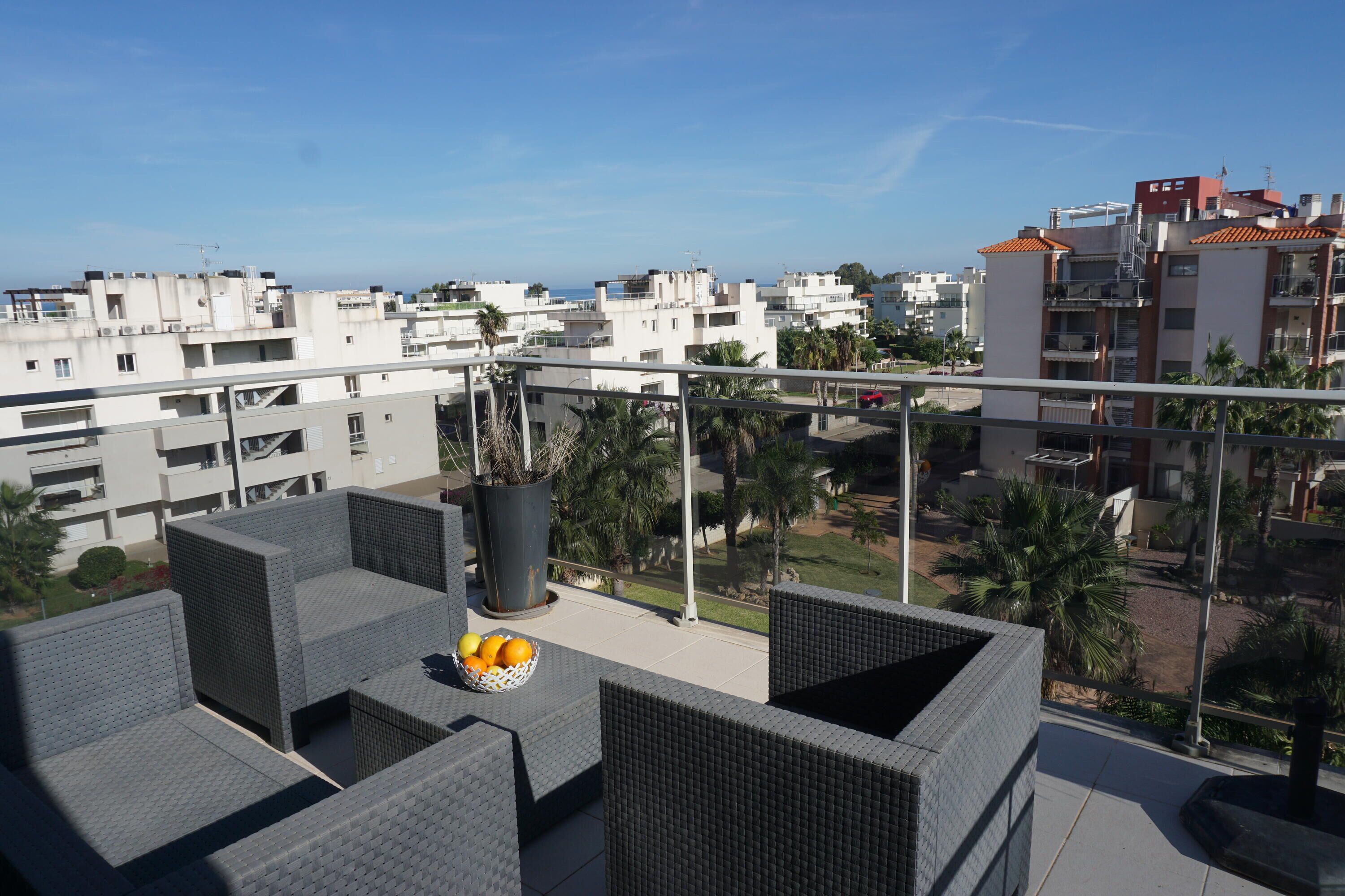 A balcony with wicker furniture and a fruit bowl, overlooking a residential area with palm trees and buildings.