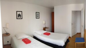 Een hotelkamer met twee bedden, een stoel, een bureau met lamp en een deur naar een andere kamer.