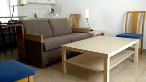 Een woonkamer met een bank, een salontafel, twee stoelen, een lamp en een keuken in de achtergrond.