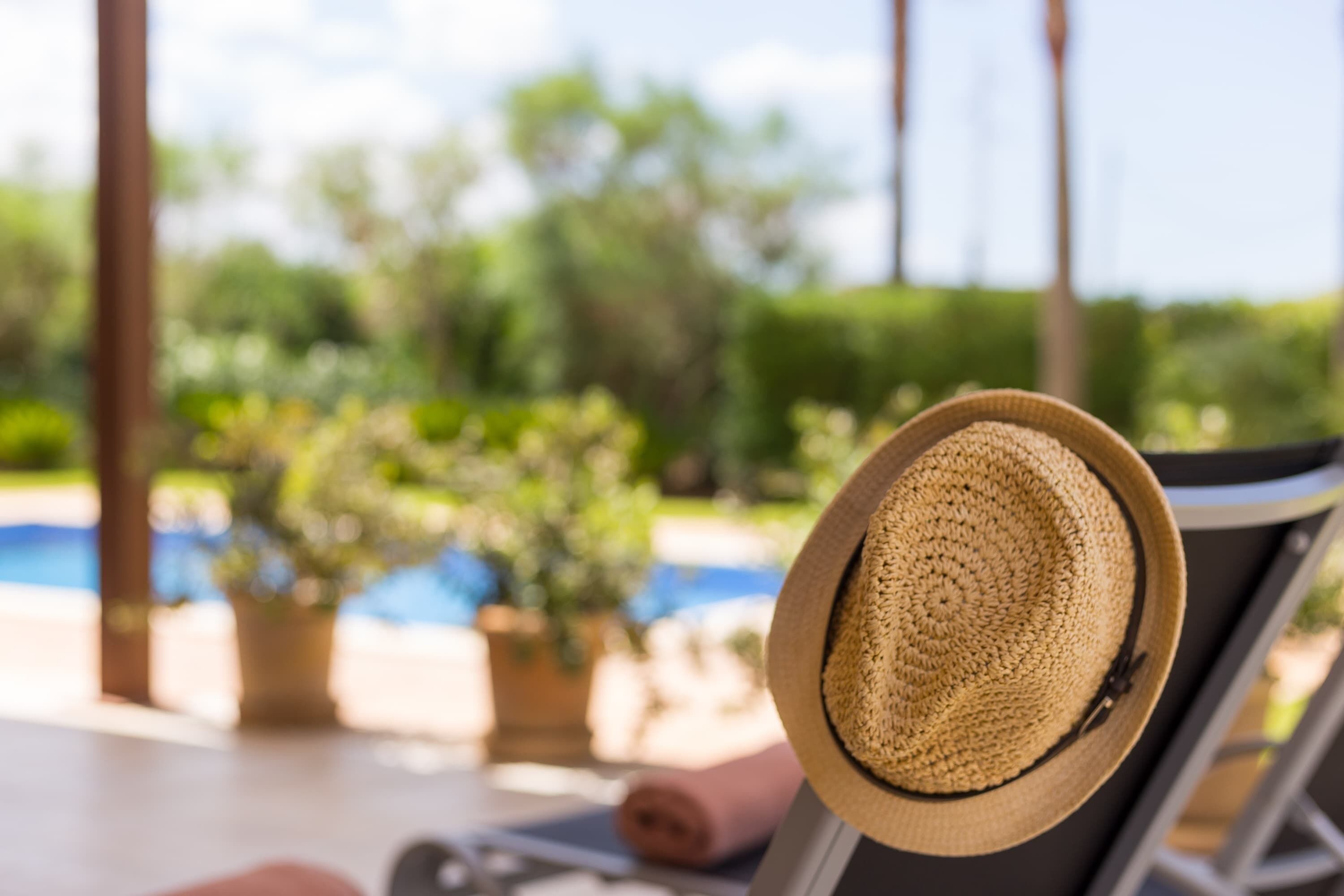 Un chapeau de paille posé sur une chaise longue près d’une piscine, avec des plantes en pot et de la verdure en arrière-plan.